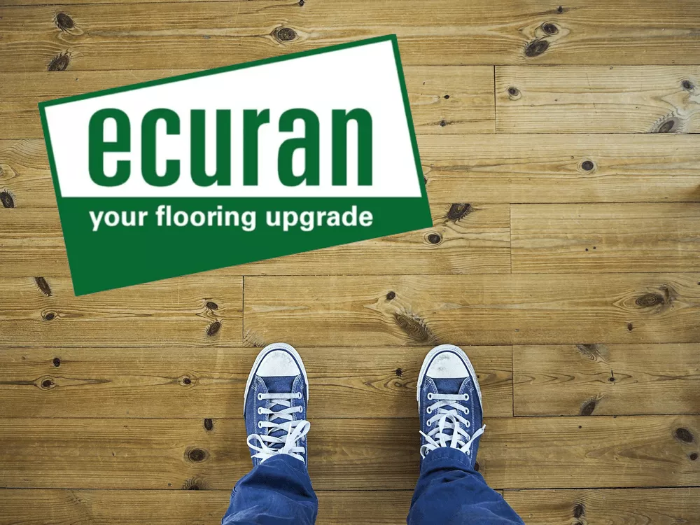 The picture shows the view from above of a pair of feet in blue shoes standing on a wooden floor covering. At the top left is the green and white ecuran logo. 