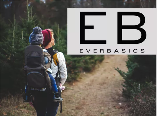 The picture shows a person with a child carrier and a child in it on a hike in the forest. The EB Everbasics logo can be seen at the top right.