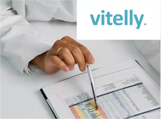 A hand with a pen and white coat points to a paper with parameters. The vitelly logo is at the top right of the picture. 